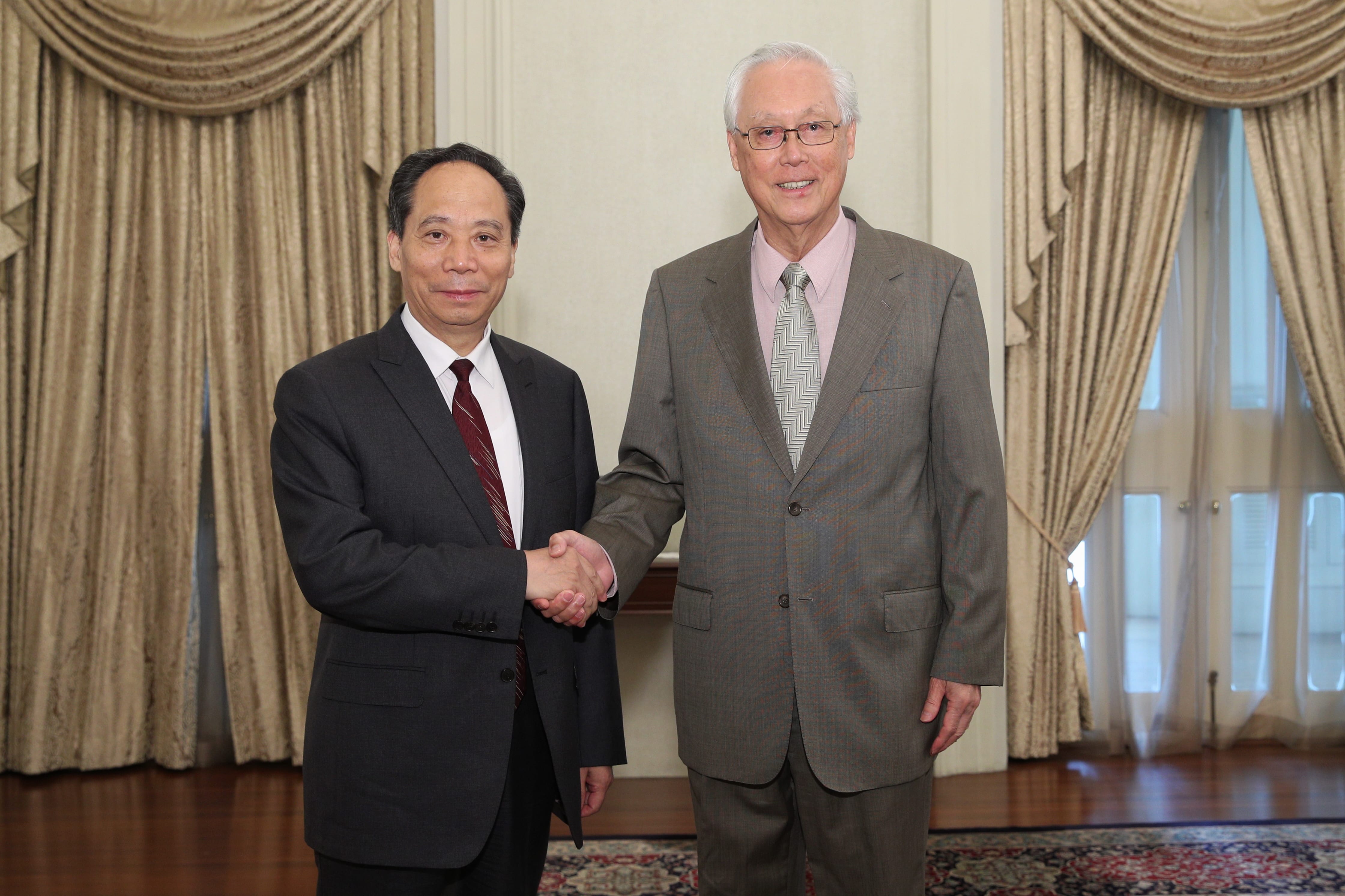 Two men in suits shaking hands indoors in front of curtains and a window.